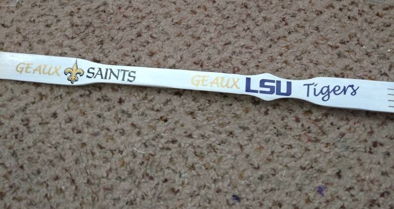 May include: A white wooden stirrer or paddle featuring the text GEAUX SAINTS with a gold fleur-de-lis emblem, followed by the text GEAUX LSU Tigers in purple and gold lettering, with a notched end visible on the right side of the object.