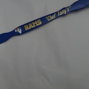 May include: A blue kitchen spatula featuring the gold text RAMS and Chef Tony's, along with a white and gold ram head logo. The handle has a contoured shape with a flat, wide end, displaying the team branding clearly in a bold, sans-serif font.