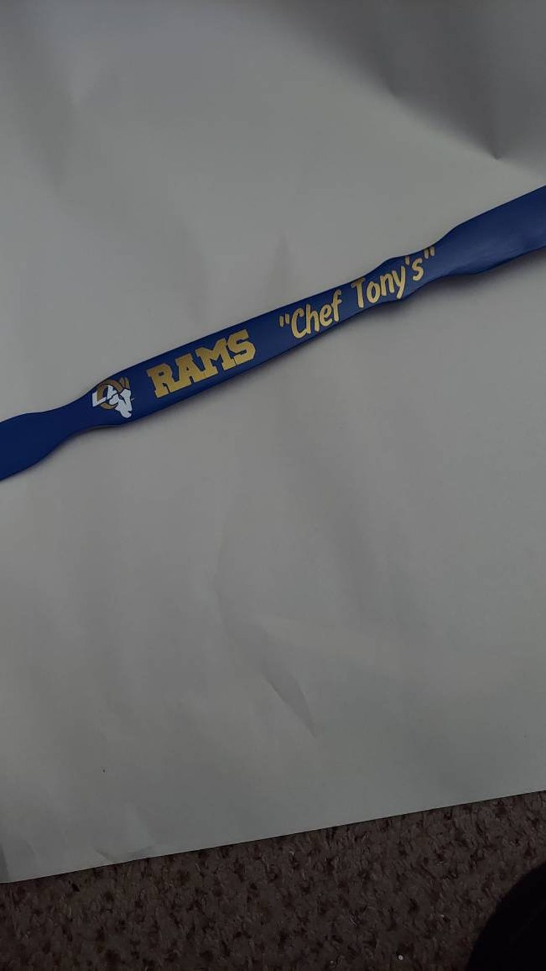 May include: A blue kitchen spatula featuring the gold text RAMS and Chef Tony's, along with a white and gold ram head logo. The handle has a contoured shape with a flat, wide end, displaying the team branding clearly in a bold, sans-serif font.