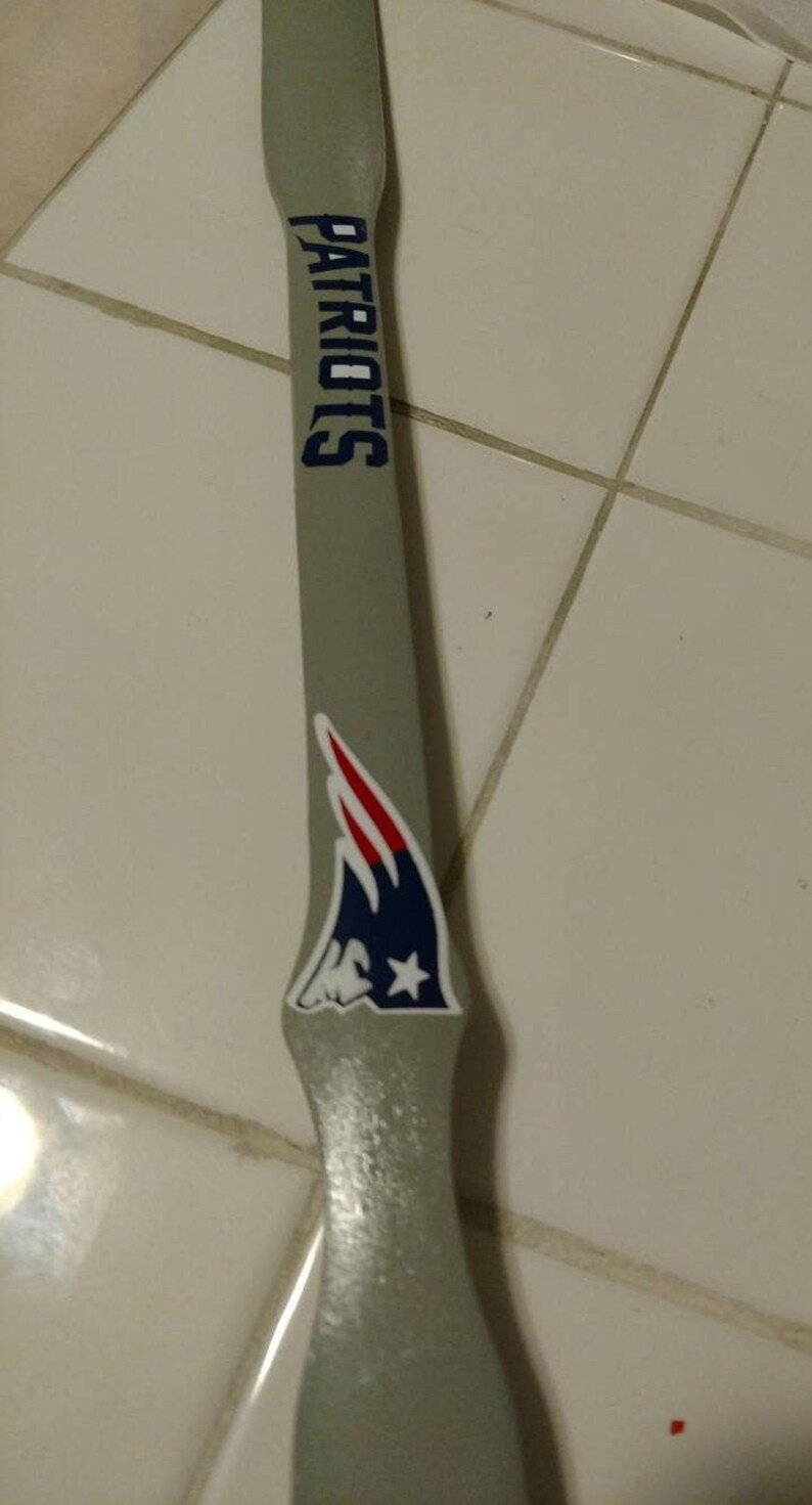 May include: A long, grey, flat wooden stirring paddle featuring the word PATRIOTS printed in dark blue capital letters and a New England Patriots team logo decal in red, white, and blue positioned below the text on the handle surface.