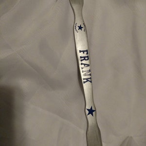 May include: A silver-toned metal shoehorn featuring the blue text FRANK printed vertically along the center. Above the name is a blue star with the text DALLAS COWBOYS curved around it, and below the name is a single blue star icon.