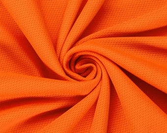 May include: A vibrant orange fabric featuring a distinct, repeating micro-mesh texture. The material is gathered into a central spiral, creating deep, flowing folds and shadows that highlight the uniform, porous pattern across the surface.
