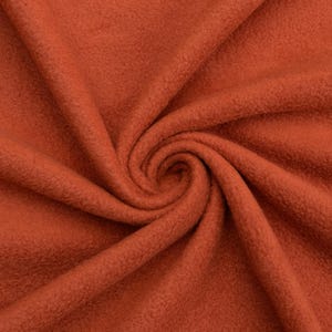 May include: Close-up of a rust-colored fleece fabric with a soft, fuzzy texture. The fabric is folded and draped, showing its drape and texture.
