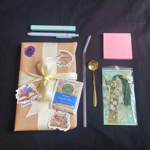 May include: A brown paper gift bag with a yellow ribbon and a variety of stickers. The stickers feature floral designs, a book, and a cup of tea. The bag also includes a box of chamomile mint tea, a gold bookmark with a floral design, a gold spoon with a flower design, a pink sticky note, and two pens.