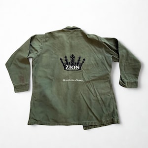 May include: An olive green jacket with long sleeves. The back of the jacket features a black crown design with the word "ZION" in white, and the phrase "The perfection of beauty" below it.