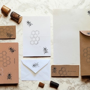 Puede incluir: Un conjunto de artículos de papelería con un diseño de abeja y panal. Incluye cuadernos, sobres, tarjetas y sellos. La paleta de colores es neutra, con elementos marrones, blancos y negros. La papelería está dispuesta sobre una superficie de mármol.