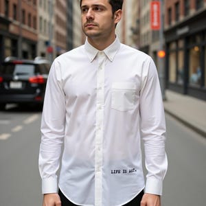 Puede incluir: Camisa blanca abotonada con bolsillo en el pecho y mangas largas. La frase "LIFE IS ART" está impresa en negro en la parte inferior. La camisa la lleva una persona en una calle de la ciudad.