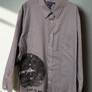 May include: A gray button-down shirt with long sleeves, a collar, and a chest pocket. The shirt features a large black and gray graphic on the lower left side with the text "LIFE IS ART." The shirt is hanging on a clear hanger.