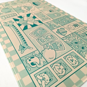May include: A rectangular, light blue and beige mat with a comic strip design featuring cats. The design includes various cat illustrations, tarot cards, and other whimsical elements. The mat has a checkered border.