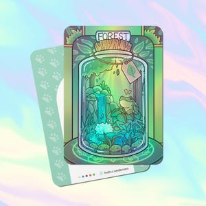May include: A glass jar with a green, blue, and white forest scene inside. The jar has a cork lid and a green leaf tag that says "FOREST" on it. A small green frog sits on a rock in the scene.
