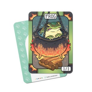 Op de afbeelding: Een fantasiekaart met een kikker in een ketel boven een vuur. De kaart is voorzien van het woord "FROG" en "1/1". De achterkant van de kaart heeft een lichtblauw pootafdrukpatroon.