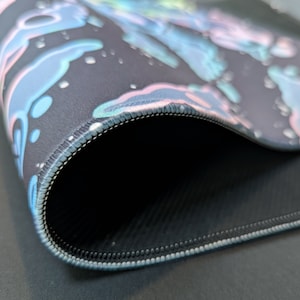 Puede incluir: Primer plano de una alfombrilla de rat&oacute;n negra con un dise&ntilde;o abstracto de colores. Los bordes est&aacute;n cosidos con hilo azul claro. La parte inferior de la alfombrilla de rat&oacute;n es negra y tiene una superficie texturizada.