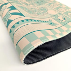 May include: A close-up of a gaming mouse pad with a detailed, colorful design. The pad features a light teal and cream color scheme with a cartoonish illustration of a dragon, skulls, and other fantasy elements. The edges are stitched with a black border.