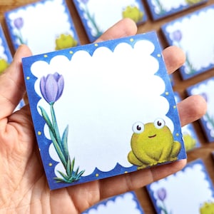 Op de afbeelding: Een set blauwe en witte notitieblok memo's met een geel kikker- en paarse tulpontwerp. Elk notitieblok heeft een gegolfde rand en gele stippen.