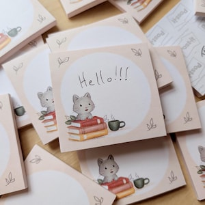 Könnte beinhalten: Quadratische Haftnotizen mit hellrosa Hintergrund. Jede Notiz zeigt eine Aquarellillustration einer grauen Katze, die auf Büchern sitzt, mit einer Kaffeetasse. Eine Notiz hat den Text "Hello!!!" in schwarzer Schrift.
