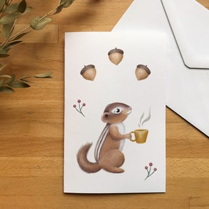 Wenskaart met een afbeelding van een eekhoorn die een warme drank drinkt, versierd met wat eikeltjes. Ontworpen in Montreal. Inclusief envelop