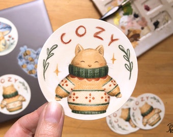 Op de afbeelding: Een ronde sticker met een witte achtergrond toont een cartoonkat die een groen-bruine trui draagt met een rood-wit patroon. Het woord "COZY" staat in rood boven de kat. De sticker is omringd door een groene, bladerrijke rand en twee gouden sterren.