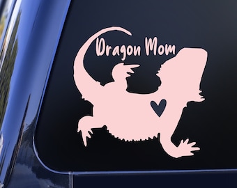 Puede incluir: Silueta rosa de un dragón barbudo con un corazón negro en una ventana de coche negro con el texto "Dragon Mom" en rosa.