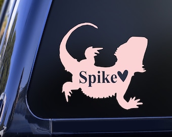 Puede incluir: Pegatina de silueta de dragón barbudo rosa con el nombre "Spike" y un pequeño corazón. La pegatina está en la ventana trasera de una camioneta negra.