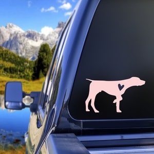 May include: Pink silhouette of a German Shorthaired Pointer dog with a small pink heart on its chest. The silhouette is on a black background, and the image is taken from the back of a truck.
