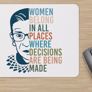 May include: A white mouse pad with a blue and white illustration of Ruth Bader Ginsburg and the text "Women belong in all places where decisions are being made".