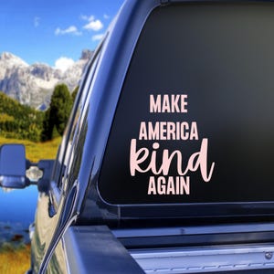 May include: A pink vinyl decal on a vehicle window. The decal reads "MAKE AMERICA kind AGAIN" in a stylized font. The background shows a mountain range and blue sky.