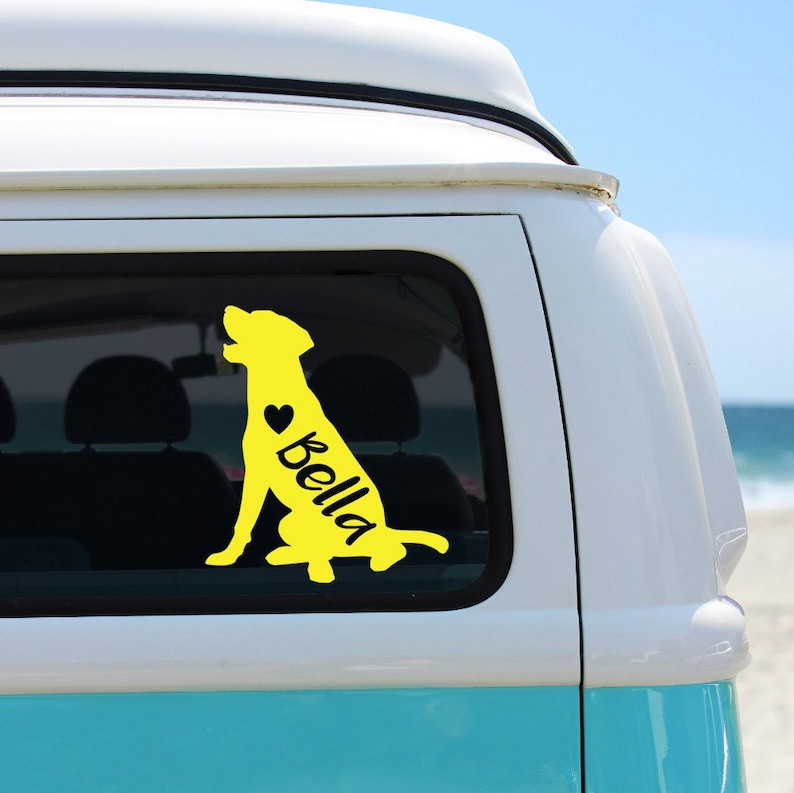 Puede incluir: Calcoman&iacute;a de vinilo amarilla con la silueta de un perro con el nombre "Bella" y un coraz&oacute;n en el lateral. La calcoman&iacute;a est&aacute; en la ventana trasera de una camioneta azul claro.
