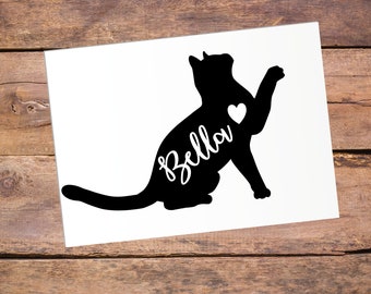 May include: Black silhouette of a cat with a heart in its chest and the name "Bella" written on its side. The cat is sitting on its hind legs with its front paw raised.