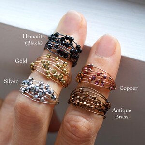 May include: Five wire wrapped rings with different colored beads. The rings are labeled Hematite (Black), Gold, Silver, Copper, and Antique Brass.
