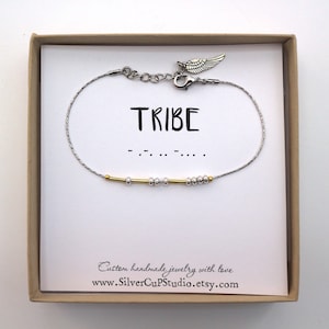 May include: A silver bracelet with a gold accent, presented in a gift box. The word "TRIBE" is printed on the card, along with Morse code. The bracelet features a small wing charm and is from SilverCupStudio.
