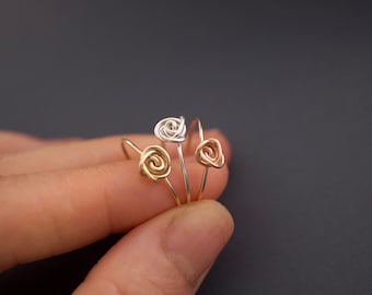 May include: Three rings with wire-wrapped rose designs in gold, silver, and rose gold. The rings are held in a hand against a dark grey background. The rings are delicate and feature a spiral design.