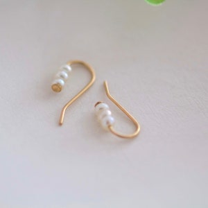 May include: Two gold-colored, curved ear cuff earrings. Each earring features a series of small, white pearls. The earrings are displayed on a white surface, with a soft, neutral background.
