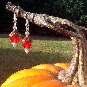 Peut inclure: Une paire de boucles d'oreilles en argent en forme de gland avec des accents de verre rouge. Les boucles d'oreilles sont suspendues à une branche avec une citrouille en arrière-plan.