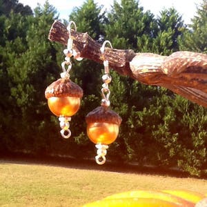 May include: A pair of silver acorn earrings with a brown and gold finish. The earrings are hanging from a branch.