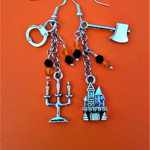 May include: A pair of silver dangle earrings with Halloween-themed charms. The charms include a house, a candlestick, handcuffs, an axe, and orange and black beads.