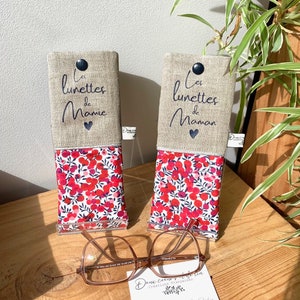 May include: Two linen eyeglass cases with red and white floral patterns. The top portion of each case is beige linen with black embroidery that reads "Les lunettes de Mamie" and "Les lunettes de Maman".