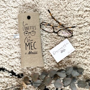 May include: A beige linen glasses case with black embroidery that reads "Les lunettes de mon mec &agrave; Moiiii". The case is lying on a white fluffy surface with a pair of brown tortoiseshell glasses and a small white business card.