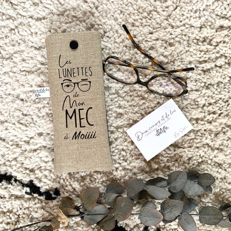May include: A beige linen glasses case with black embroidery that reads "Les lunettes de mon mec &agrave; Moiiii". The case is lying on a white fluffy surface with a pair of brown tortoiseshell glasses and a small white business card.
