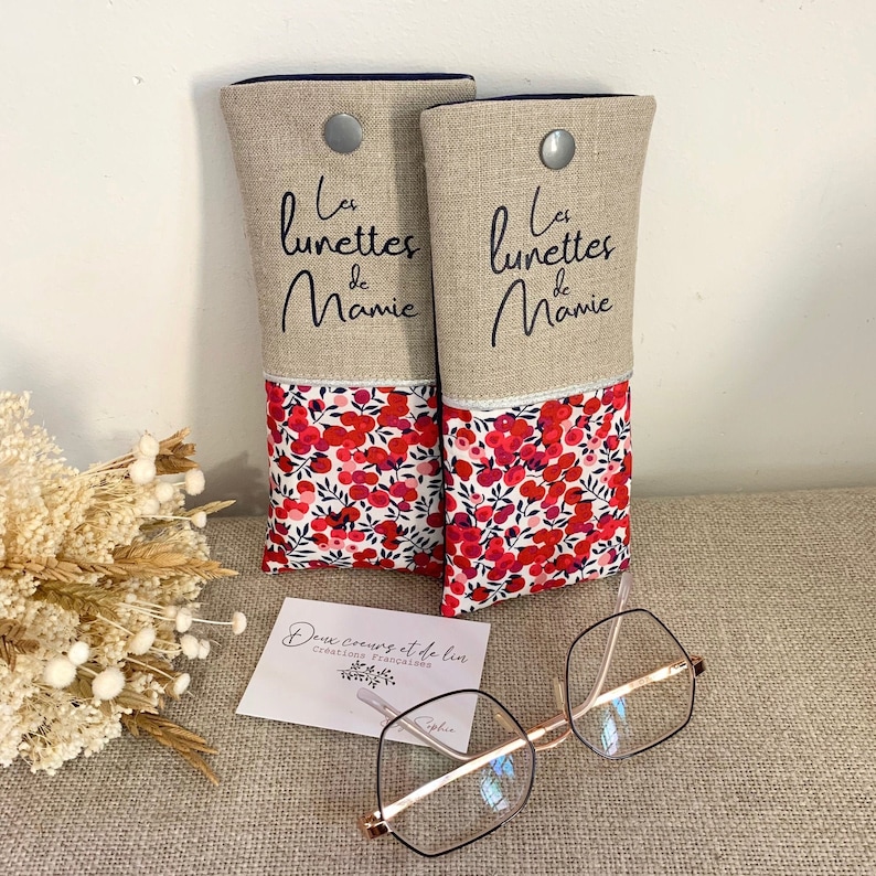 May include: Two linen eyeglass cases with a red floral pattern and the text "Les lunettes de Mamie" in black. The cases are sitting on a beige surface with a business card that says "Deux coeurs et du lin creations francaises".