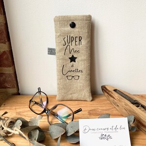 May include: A beige linen glasses case with black text that reads "SUPER MEC &agrave; Lunettes" and a black star. The case has a black button closure.