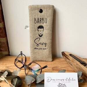 May include: A beige linen glasses case with a black button closure. The case has a black graphic of a man with a beard and glasses and the text "Barbu Sexy Mais pas que!" printed on it.