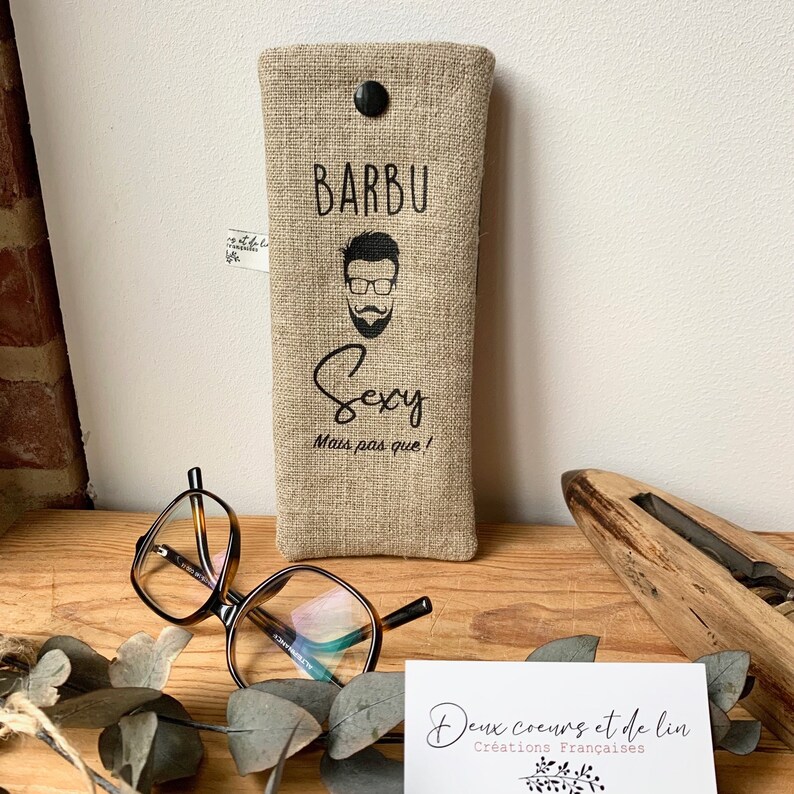 May include: A beige linen glasses case with a black button closure. The case has a black graphic of a man with a beard and glasses and the text "Barbu Sexy Mais pas que!" printed on it.