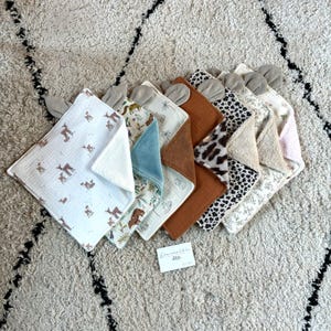 Può includere: Una collezione di bavaglini per bambini in vari modelli e colori. I panni presentano stampe animalier, disegni floreali e colori a tinta unita. Ogni panno ha una superficie morbida e strutturata ed è sormontato da piccoli accenti grigi a forma di orecchio.