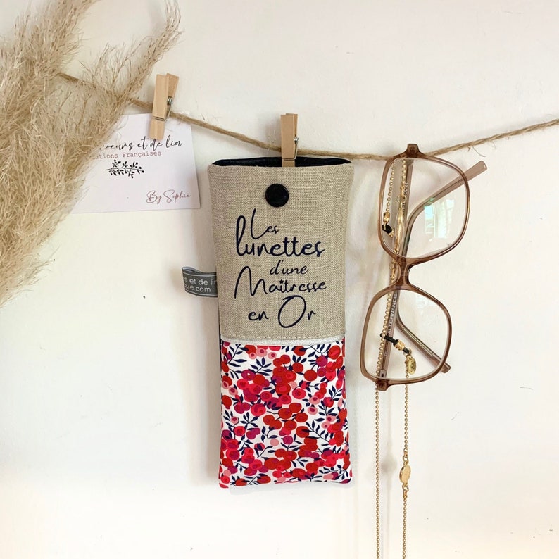 May include: A beige linen glasses case with a floral print lining. The case has a black button and the text "Les lunettes d'une Maitresse en Or" is embroidered on the front.