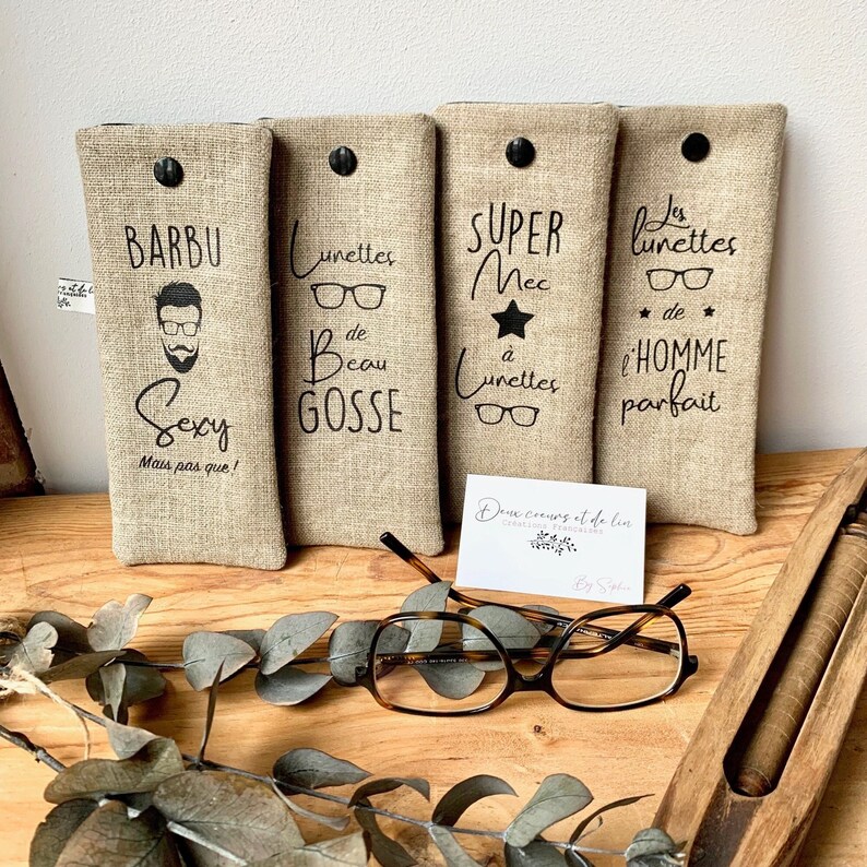 May include: Three beige linen eyeglass cases with black text and a black button closure. The cases have different sayings on them: "Barbu Sexy Mais pas que!"  "Lunettes de Beau Gosse" and "Super Mec a Lunettes" and "Les Lunettes de l'Homme Parfait".