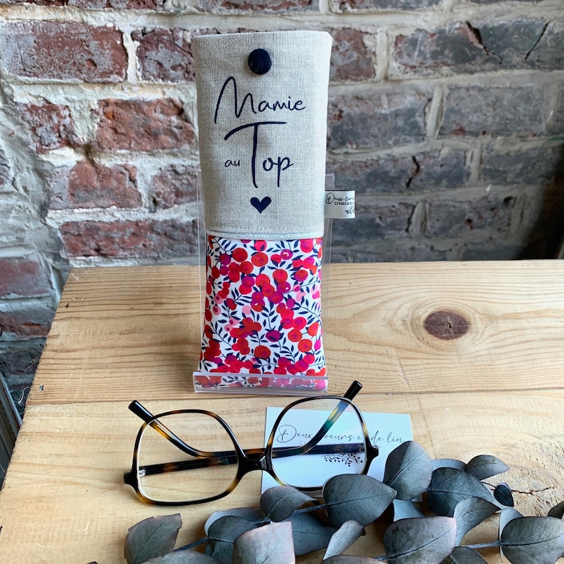 May include: A linen eyeglass case with a red and white floral pattern. The case has a flap with the text "Mamie au Top" and a small heart.