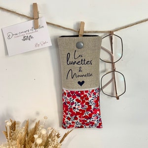 May include: A beige linen glasses case with a floral print lining. The case has a black button closure and the text "Les lunettes de Mounette" is printed on the front.
