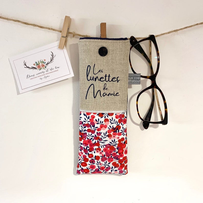 May include: A beige linen glasses case with a floral fabric lining. The case has a button closure and the text "Les lunettes de Mamie" is embroidered on the front.