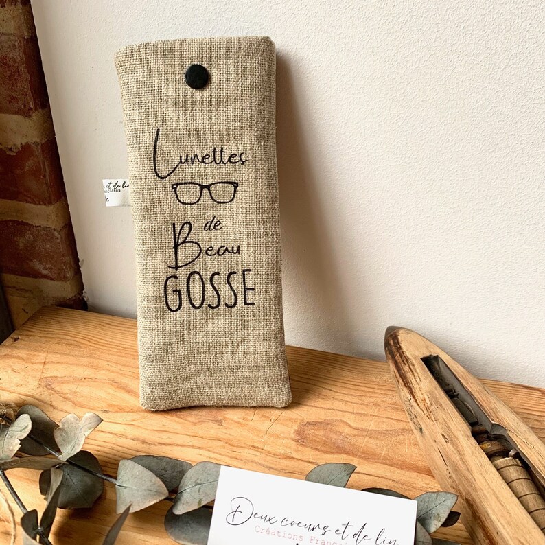 May include: A beige linen glasses case with black stitching and a black button closure. The case has the text "Lunettes de Beau Gosse" embroidered on it.