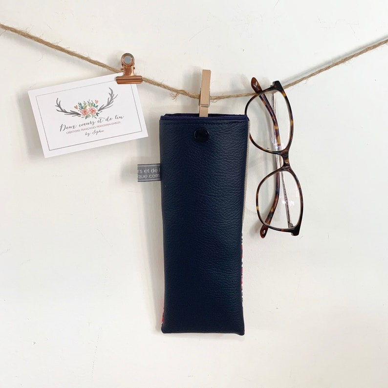 May include: A navy blue glasses case with a zipper closure. The case is hanging from a string with a wooden clothespin. The case is decorated with a red and white floral pattern. A pair of tortoiseshell glasses is hanging from the string.